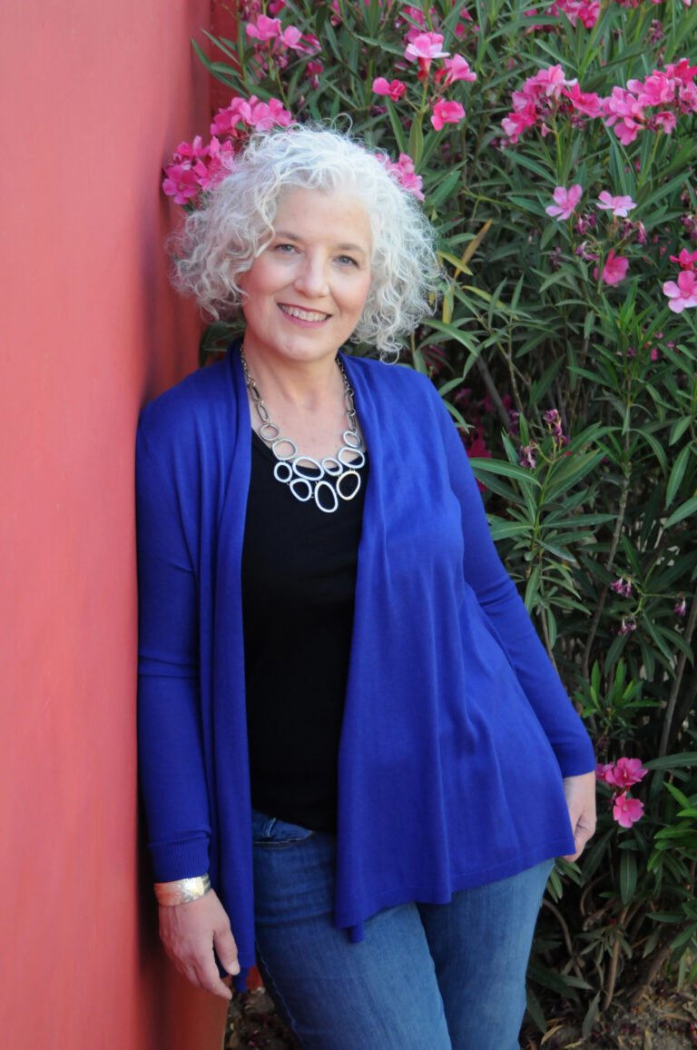Outdoor portrait by Laura Voight: woman with white curly hair in a blue cardigan beside a pink wall